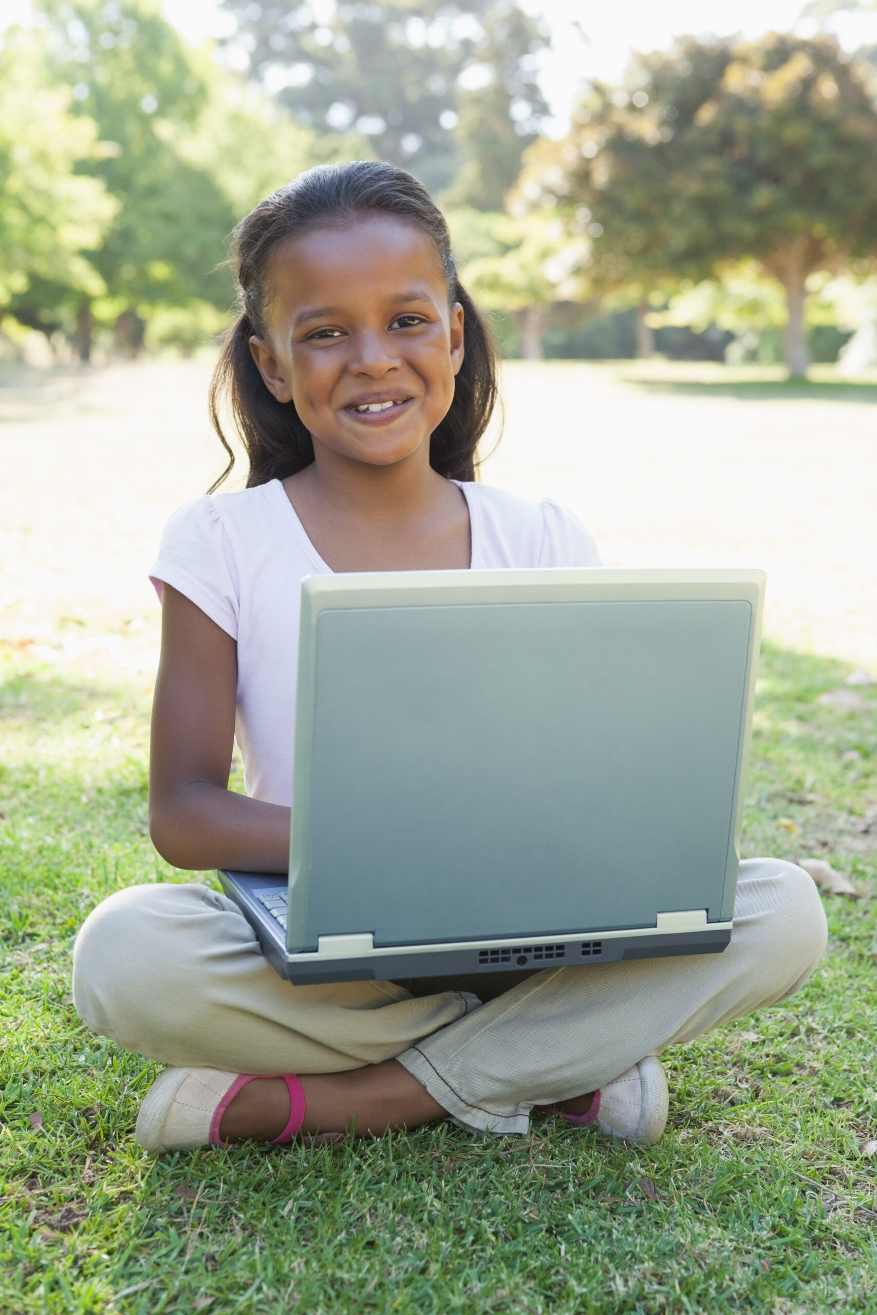 Little girl sitting on grass using laptop smiling at camera on a sunny day